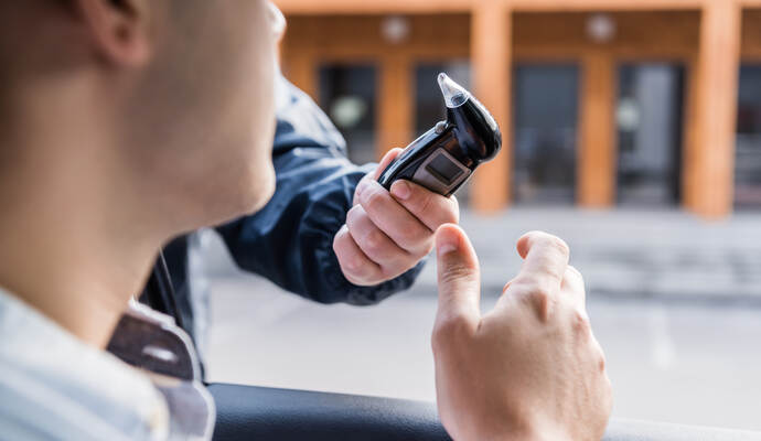 Der 55-jährige VW-Fahrer widersetzte sich einer ersten Verkehrskontrolle in Ötisheim. Erst durch Unterstützung einer Streife vom Polizeirevier Bretten konnte der stark alkoholisierte Mann bei einem Steinbruch zwischen Ötisheim und Enzberg gestoppt werden. cropped view of policeman giving breathalyzer to driver in car, blurred foreground.