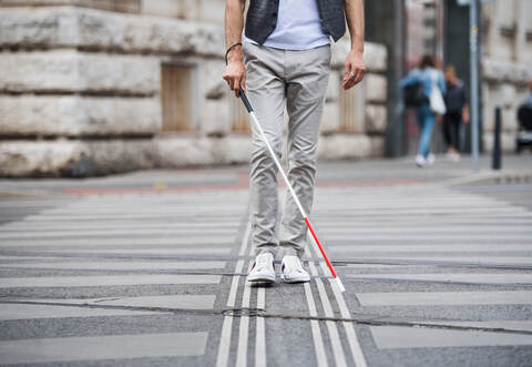 Midsection of young blind man with white cane walking across the street in city.