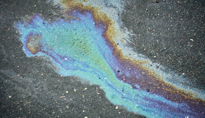 Weil Öl und Schmierstoffe ausgelaufen sind, muss die Fahrbahn erneuert werden. A vibrant oil spill spreads across the dark asphalt, revealing shades of blue, purple, and green. The unique patterns draw atten