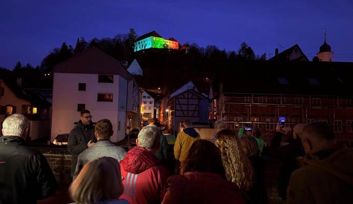 Begeisterte Schaulustige betrachten die bunt beleuchtete Fassade des Neuenbürger Schlosses. Die Farben können interaktiv über eine Website und einen QR-Code ausgewählt werden.