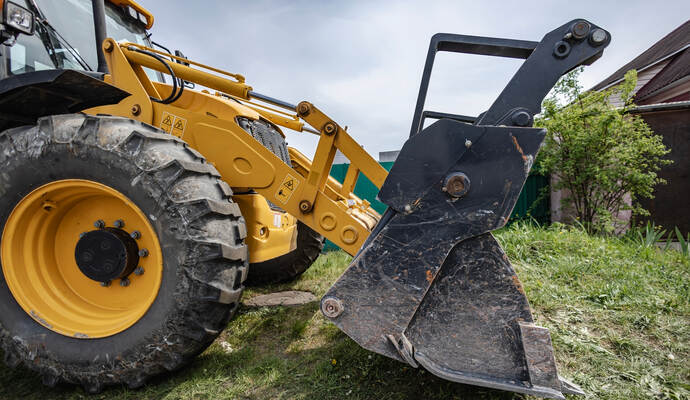 A loader is parked in a yard, ready to dig soil and lift materials. The machine sits near a house with green grass around it on 