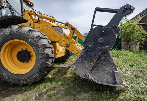 A loader is parked in a yard, ready to dig soil and lift materials. The machine sits near a house with green grass around it on 