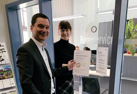 Bürgermeister Klaus Mack hängt mit seiner Sekretärin Edith König das erste Hinweisplakat am Bürgerservice im Rathaus auf.