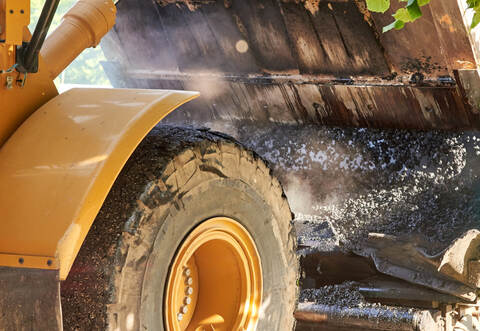 Bucket tractor pours hot asphalt into a grader, construction of a new road, material for road construction close-up.