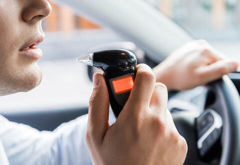 partial view of man blowing into breathalyzer while sitting in car, blurred background.