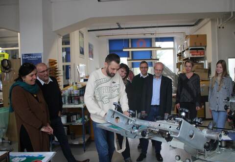 Schauen Mark G., Teilnehmer im Projekt Andante, bei der Arbeit an der Siebdruckmaschine über die Schulter: Ute Hötzer (Geschäftsführerin von Q-Prints &amp; Service), Harald Stickel (Geschäftsführer Plan B), Susanne Stiegel (Beratungsstellenleitung Diakonie Suchthilfe), Hartmut Schölch, Achim Oeder und Miriam Mohr vom Jobcenter Enzkreis sowie Projektmitarbeiterin Natalie Richter (von links).