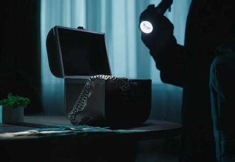 A thief commits a burglary, stealing valuables in a dark apartment. A jewelry box illuminated by a beam from a lantern in the ha