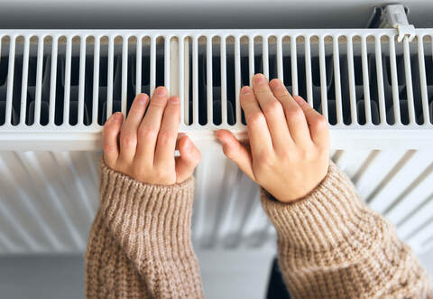 Child wearing brown woolly jacket warming cold hand in front of heating radiator in winter time. Electric or gas heater at home.